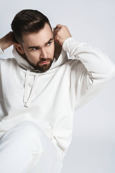 Handsome man wearing blank white hoodie and pants sitting on light gray background - Fotoğraf, Görsel