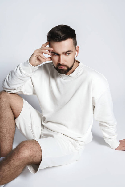 Handsome man wearing white sweatshirt and shorts with wireless earbuds sitting against gray background - Foto, imagen
