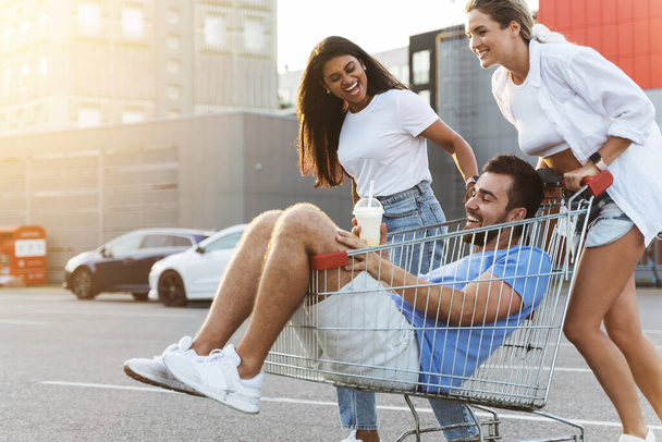 Three diverse friends having fun and riding shopping cart on parking lot near supermarket during summer evening - Foto, Bild