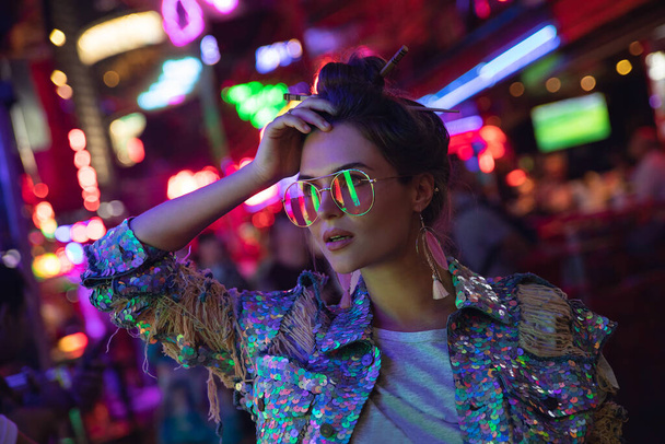 Young stylish woman wearing jacket with shining sequins on the city street with neon lights - Фото, изображение