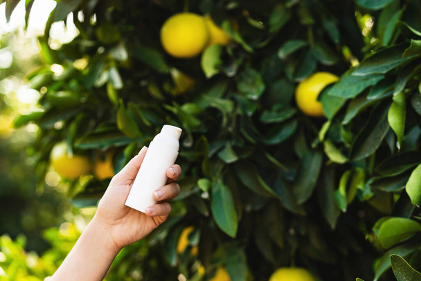 Woman holds a white bottle of skin care product in her hand on blurry lemon tree background. - Photo, Image