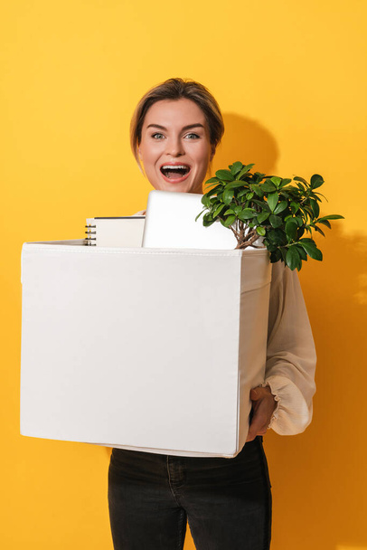 jong gelukkig vrouw holding box met persoonlijke items na job promotie tegen geel achtergrond - Foto, afbeelding