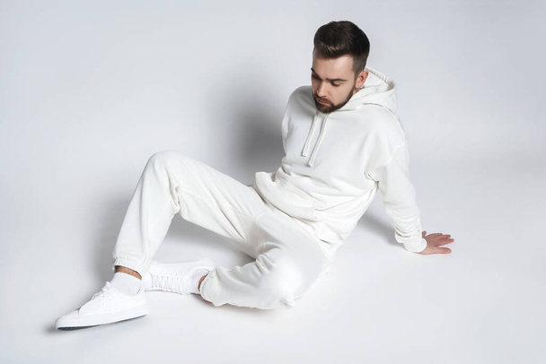 Handsome man wearing blank white hoodie and pants sitting on light gray background - Фото, зображення