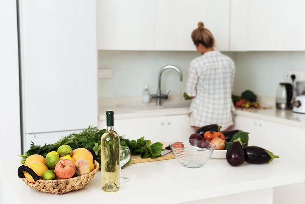 Bottle of white wine, basket of fruits and different vegetables on white kitchen table - Valokuva, kuva