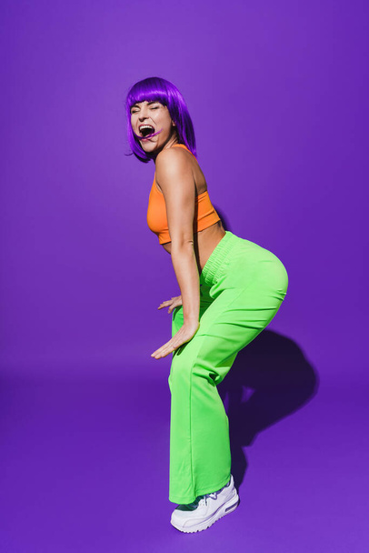 Carefree woman dancer wearing colorful sportswear twerking against purple background - 写真・画像