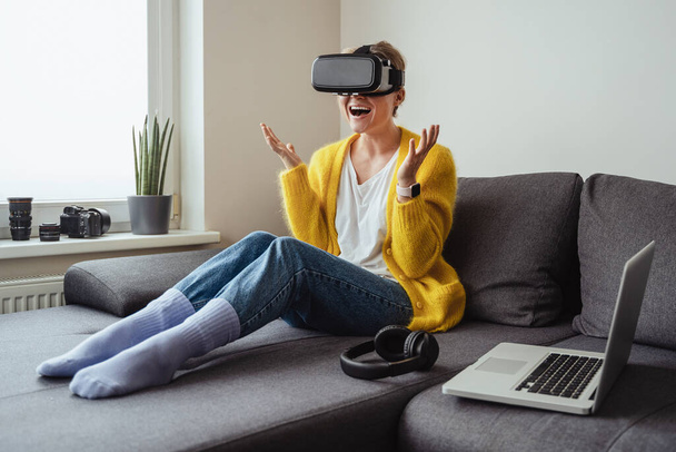 Excited young woman sitting on the sofa and using virtual reality headset at home - Foto, afbeelding