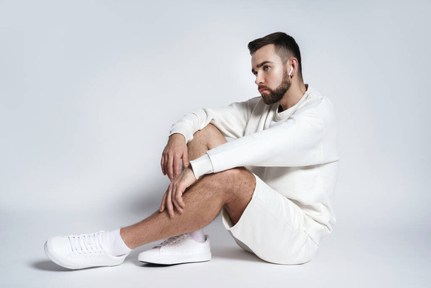 Handsome man wearing white sweatshirt and shorts with wireless earbuds sitting against gray background - Foto, Imagen