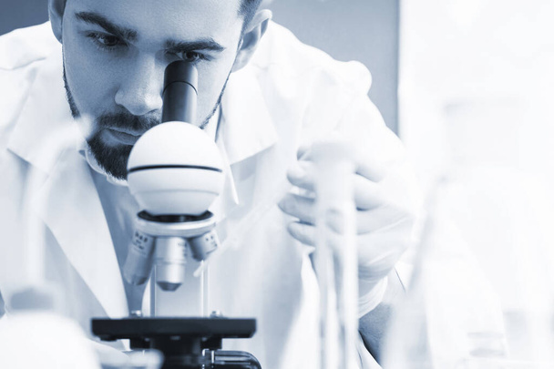 Man scientist is using microscope in a laboratory during science research work. Tinted in blue monochrome colors for scientific look. - Photo, Image