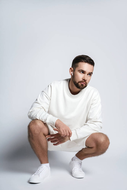 Handsome man wearing white sweatshirt and shorts with wireless earbuds sitting against gray background - Fotoğraf, Görsel
