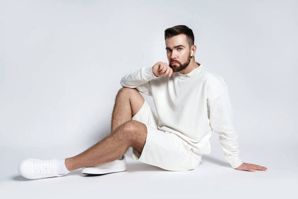 Handsome man wearing white sweatshirt and shorts with wireless earbuds sitting against gray background - Fotoğraf, Görsel