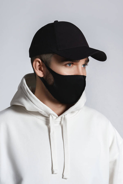Young man wearing white hoodie, black baseball cap and cloth face mask against gray background - Foto, afbeelding