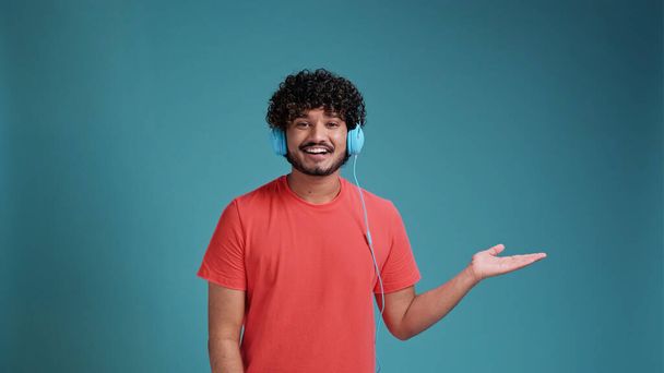 latin spanish man listening to music using headphones in coral t-shirt on blue studio background. pointing with hand to the side - 写真・画像