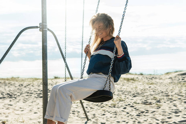 Young pretty woman is swinging on the swing and smiling at the sandy beach on a sunny day. - Fotoğraf, Görsel