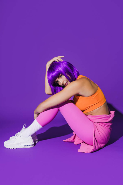 Portrait of young woman wearing colorful sportswear sitting against purple background - Photo, image