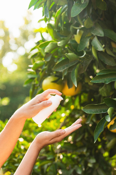 Woman holds a white bottle of skin care product in her hands on blurry lemon tree background. - Valokuva, kuva