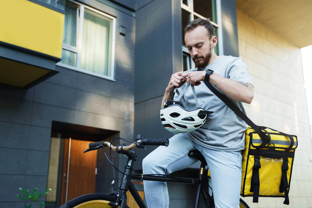 Young express delivery courier with insulated bag on his back puts on a bike helmet while sittitng on his bicycle. - Foto, Bild