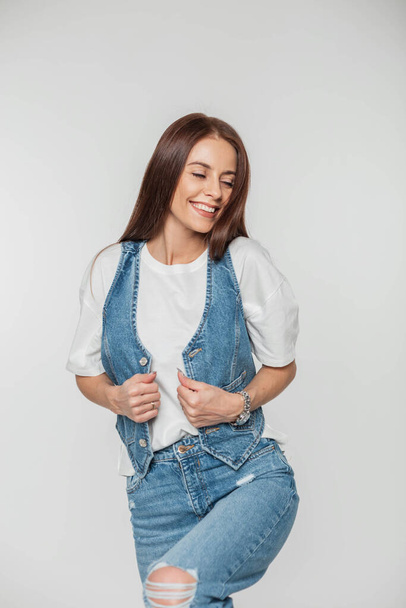 Mooi vrolijk schattig jong vrouw model met een leuke glimlach in trendy mode denim kleding met een jeans vest, t-shirt en jeans poseren op een witte achtergrond in de studio - Foto, afbeelding