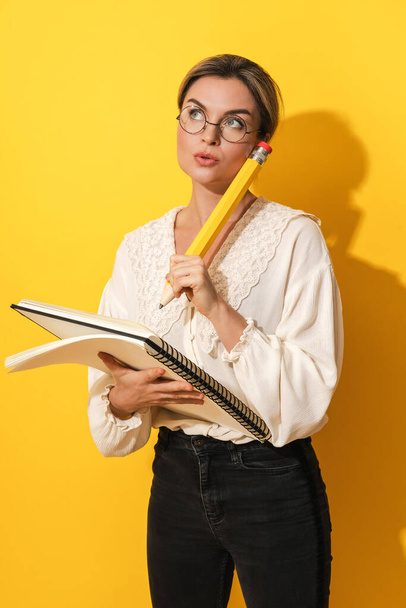 Thoughtful young woman wearing glasses holding big pencil and notebook on yellow background - Fotó, kép