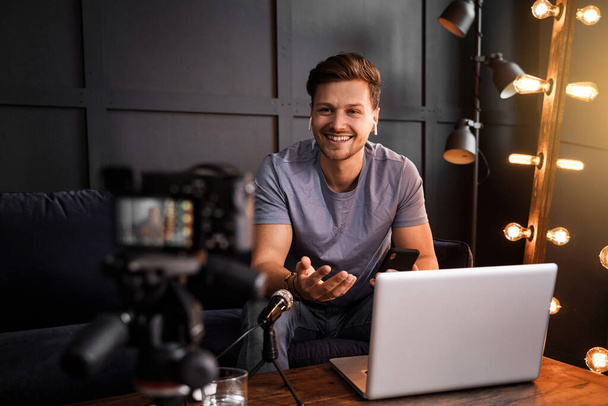 Kanepede oturan genç sakallı blogcu kameraya bakıyor ve loş bir odada dizüstü bilgisayarın önünde gülümsüyor.. - Fotoğraf, Görsel