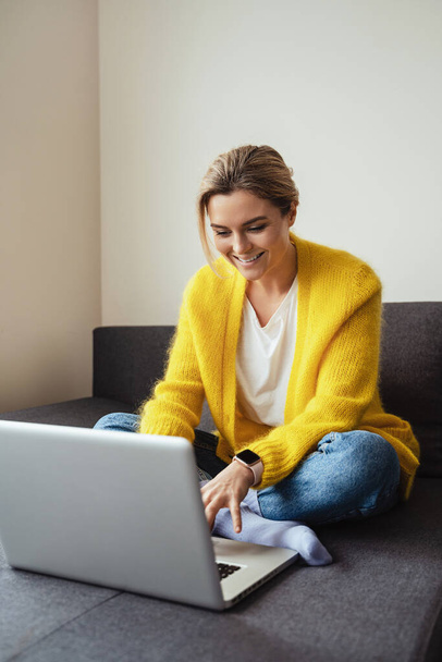 Young woman wearing yellow cardigan sitting on the sofa and using laptop computer - Фото, изображение