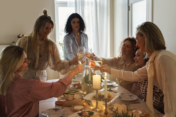 Young happy women friends clinking glasses and smiling at the festive table served with food and white wine, decorated with candles. - Photo, Image