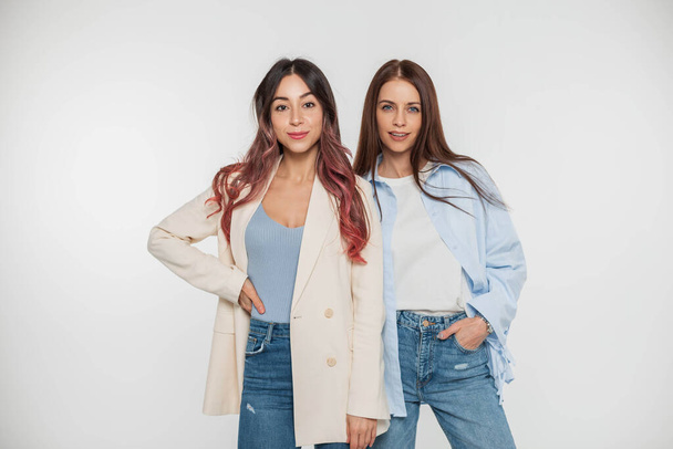 Две модные красивые девушки девушки с улыбками в модной повседневной джинсовой одежде позируют на белом фоне в студии. Красота и стиль - Фото, изображение