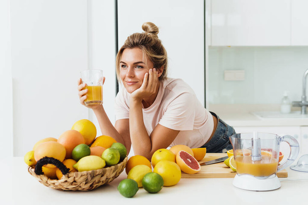 Young woman drinking freshly squeezed homemade orange juice in white kitchen - Valokuva, kuva