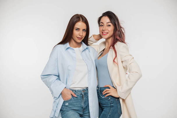 Дві модні красиві подруги дівчат в стильному повсякденному одязі з сорочкою, зверху і блейзером з джинсами позують і розважаються на білому тлі
 - Фото, зображення