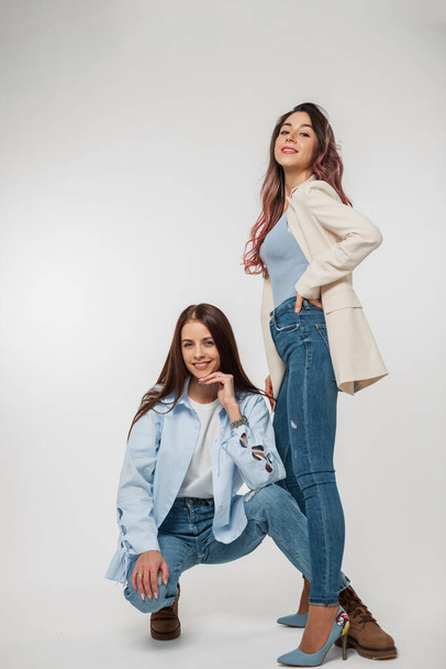 Zwei modische hübsche Freundinnen im Jeans-stylischen Casual-Outfit sitzen und posieren auf weißem Hintergrund im Studio - Foto, Bild