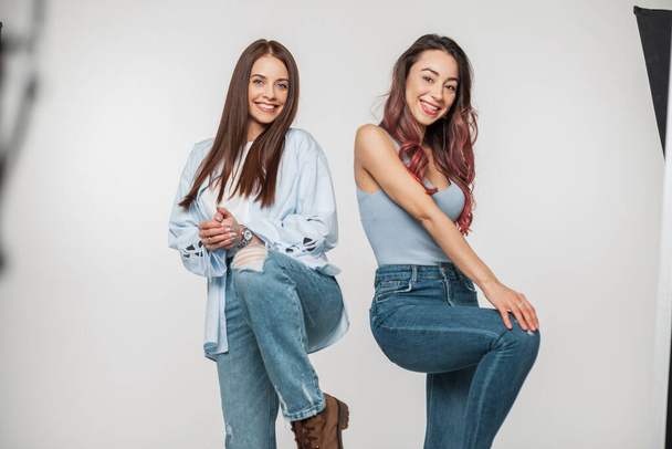 Dvě legrační krásné veselé dívky v módní džínové ležérní oblečení představuje a bavte se na focení na bílém pozadí ve studiu - Fotografie, Obrázek