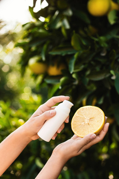 Woman holds a white bottle of skin care product and half of a juicy lemon in her hands on blurry lemon tree background. - Valokuva, kuva