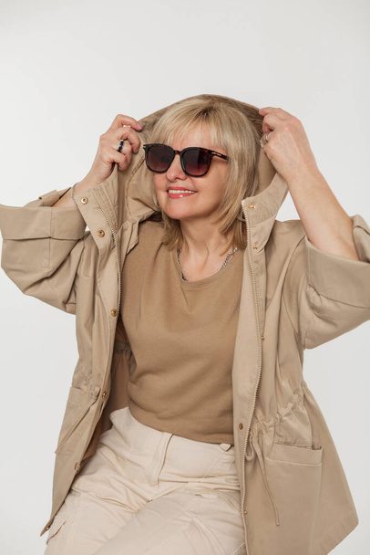 Модная красивая пожилая женщина в старинных солнцезащитных очках и стильной бежевой куртке надевает капюшон на белом фоне в студии - Фото, изображение