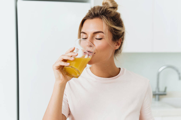 Young woman drinking freshly squeezed homemade orange juice in white kitchen - Photo, Image
