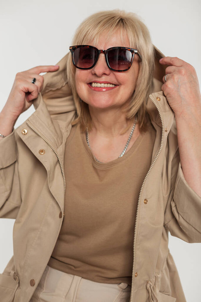Happy elderly blonde pretty aged woman with a smile with vintage sunglasses in fashionable beige clothes with a windbreaker jacket puts on a hood on a white background - Фото, зображення