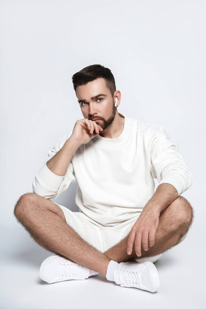 Handsome man wearing white sweatshirt and shorts with wireless earbuds sitting against gray background - Φωτογραφία, εικόνα