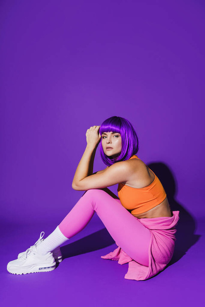 Portrait of young woman wearing colorful sportswear sitting against purple background - Foto, immagini