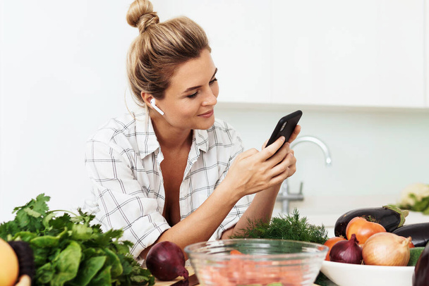 Молода красива жінка з бездротовими навушниками використовує смартфон під час приготування їжі на сучасній білій кухні
 - Фото, зображення