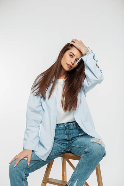 Стильная красивая молодая женщина модель в модной джинсовой одежде в рубашке и джинсах сидит на стуле и выпрямляет волосы на белом фоне - Фото, изображение