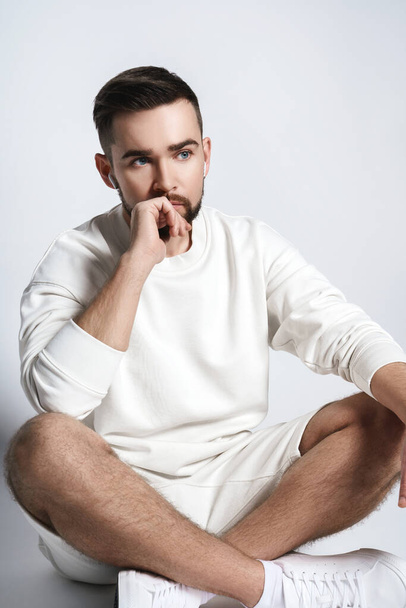 Handsome man wearing white sweatshirt and shorts with wireless earbuds sitting against gray background - Fotografie, Obrázek