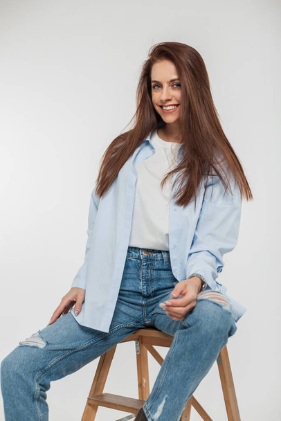Happy beautiful girl with long hair in casual fashion clothes with a shirt, t-shirt and jeans sits and smiles on a white background - Φωτογραφία, εικόνα