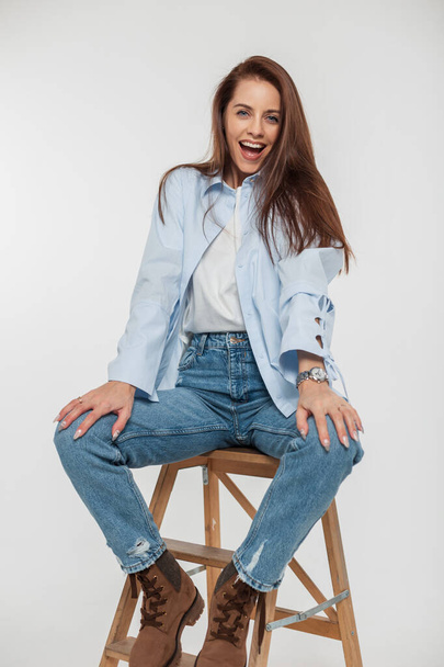 Glückliche hübsche junge Frau Modell mit positiven Emotionen in modischen Jeans Casual Outfit sitzen und Spaß haben auf einem Stuhl auf weißem Hintergrund - Foto, Bild