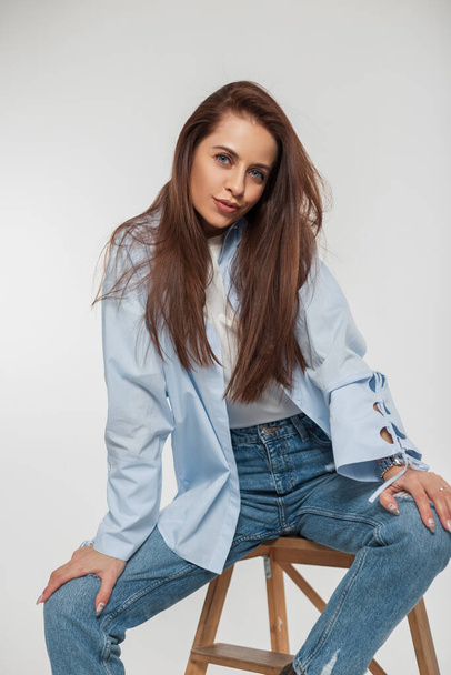 Cheerful beautiful young woman model with long hair in fashion clothes with a shirt and jeans sits on a wooden chair on a white background - Foto, Bild