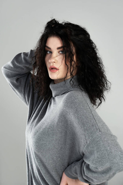 Portrait of young and sexy woman with black wavy hair wearing turtleneck jumper posing on gray background in studio - Foto, Imagem