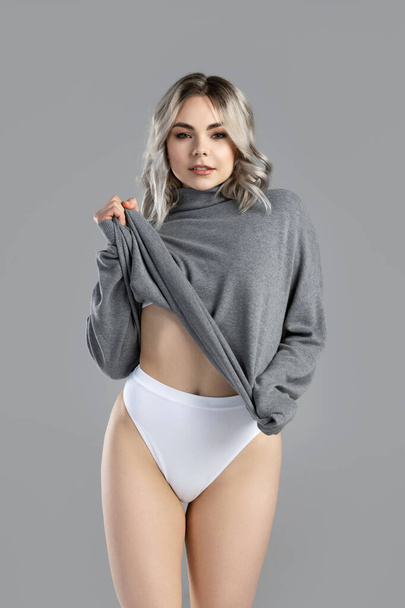 Portrait of young and sexy woman with blond hair wearing turtleneck jumper is posing on gray background in studio - Valokuva, kuva