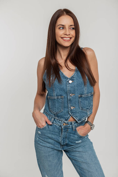 Happy beautiful young woman with a sweet smile with a slim body in fashionable retro denim clothes with jeans and a vest on a white background. Vintage denim fashion and style - Foto, afbeelding