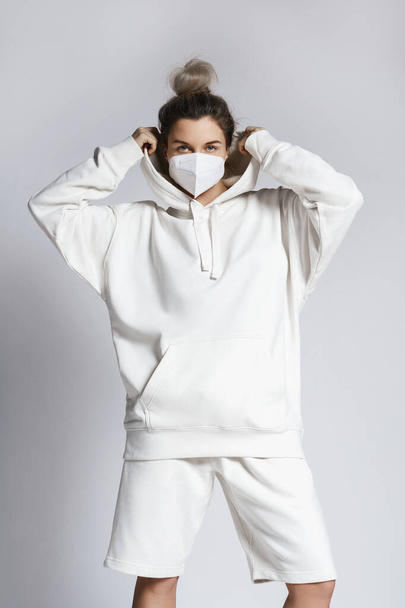 Young woman wearing white hoodie and ffp2 respirator mask against gray background - Valokuva, kuva