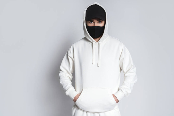 Young man wearing white hoodie, black baseball cap and cloth face mask against gray background - Фото, изображение