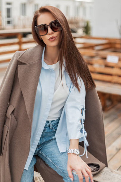 Elegante mooie blanke yin model meisje met vintage bril in trendy casual kleding met een jas, shirt, t-shirt en jeans zit en heeft plezier op straat in de stad - Foto, afbeelding
