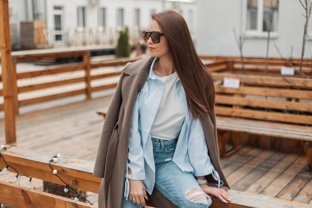 Bella ragazza alla moda con occhiali da sole vintage in un vestito alla moda casual con un cappotto elegante, camicia e jeans si siede e si riposa su una recinzione di legno sulla strada - Foto, immagini