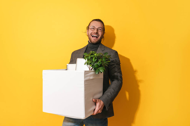 Young happy man holding box with personal items after job promotion against yellow background - Valokuva, kuva